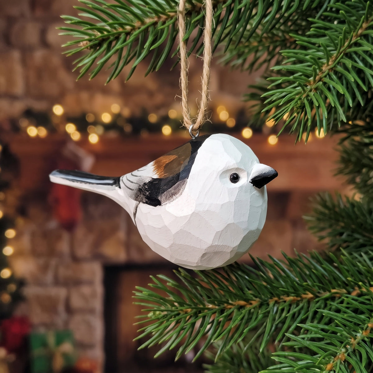A faceted wooden bird ornament, shaped like a small white bird with black, brown, and orange details, hanging from a natural twine loop on a green Christmas tree branch in front of a warm, blurred holiday fireplace background.