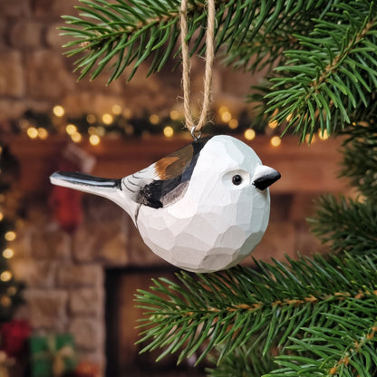 A faceted wooden bird ornament, shaped like a small white bird with black, brown, and orange details, hanging from a natural twine loop on a green Christmas tree branch in front of a warm, blurred holiday fireplace background.