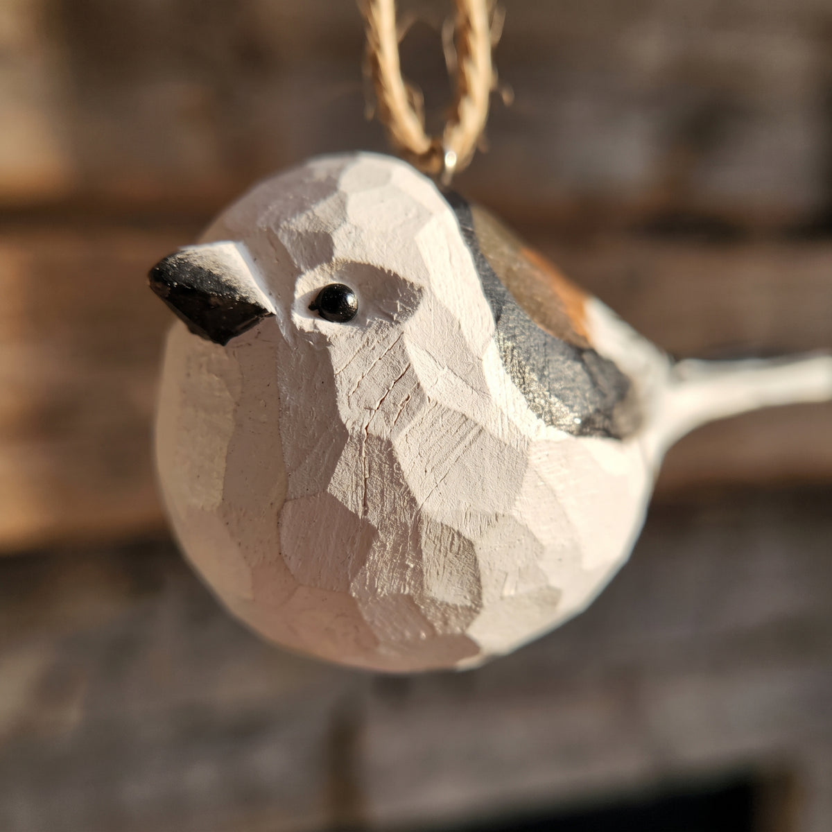 A close-up of a hand-carved wooden Long-tailed tit ornament, likely a chickadee, with a geometric polygonal pattern and a natural twine hanger, set against a blurred rustic wooden background.