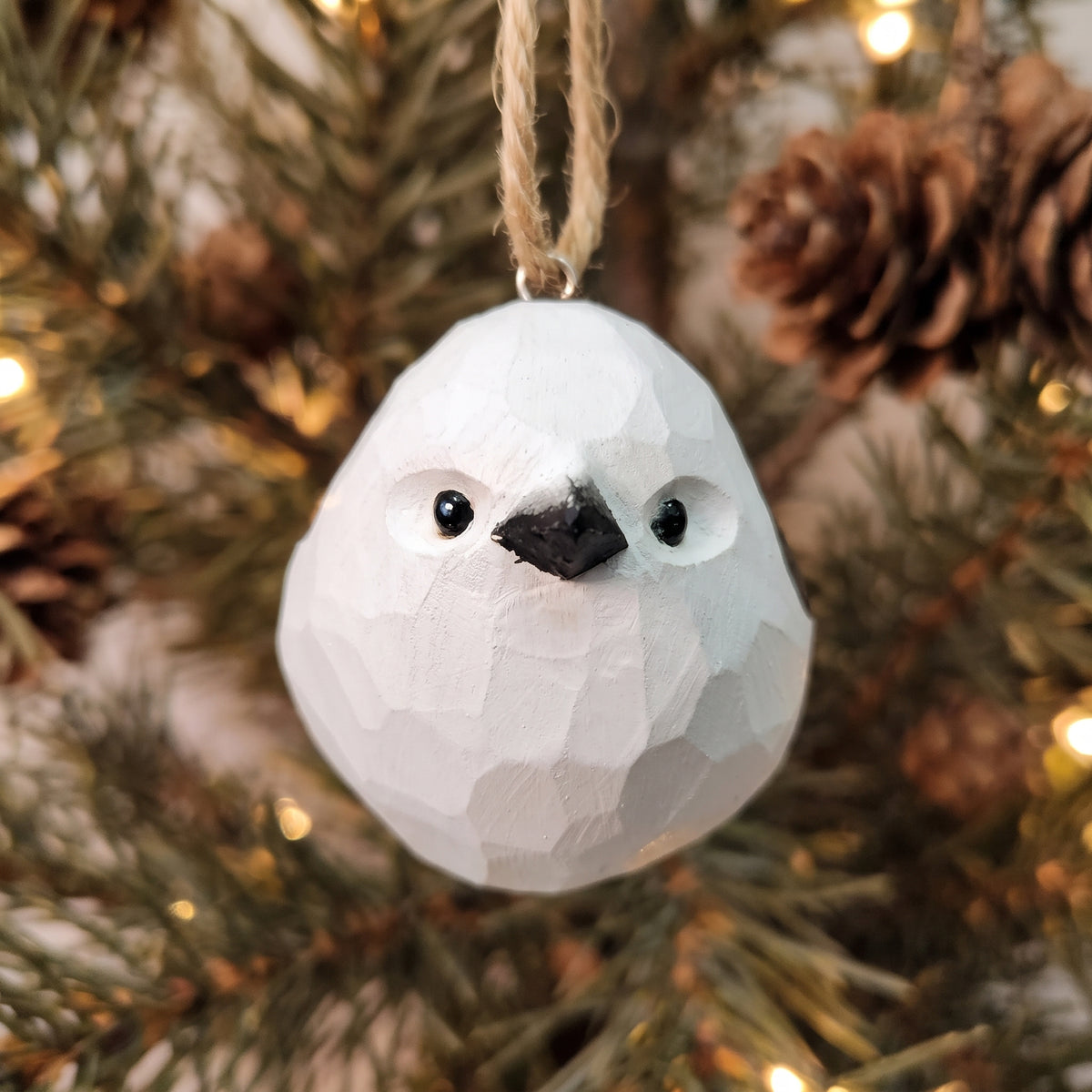 A white painted Long-tailed tit ornament with a black beak and black eyes, hanging from a jute string against a blurred Christmas tree background with pine needles and warm lights.