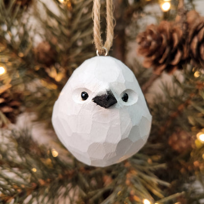 A white painted Long-tailed tit ornament with a black beak and black eyes, hanging from a jute string against a blurred Christmas tree background with pine needles and warm lights.