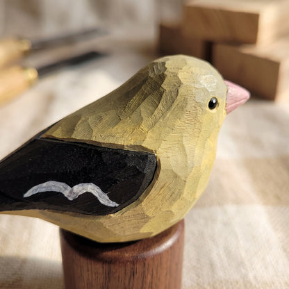 A hand-carved wooden Female American Goldfinch figurine with a yellow-green body, black wing, pink beak, and small white accent, mounted on a wooden stand.