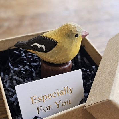 A small yellow Female American Goldfinch figurine with black wings and a pink beak sits in a brown kraft gift box filled with black shredded paper and a card that reads 'Especially For You'. The box is placed on a wooden table.