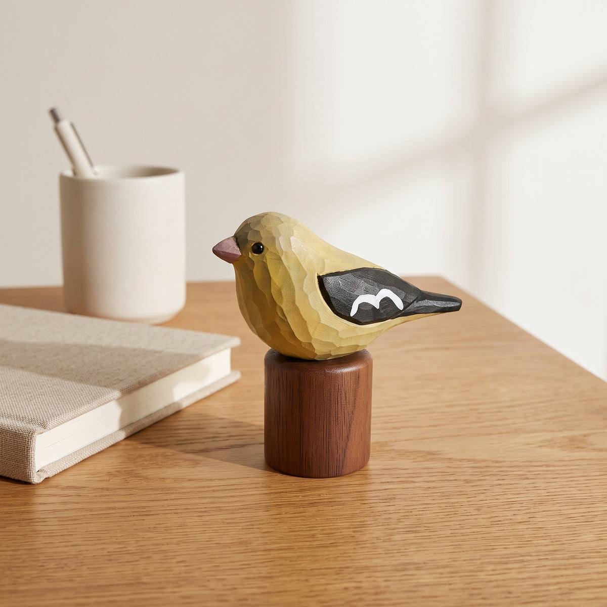 A handcrafted wooden Female American Goldfinch figurine in yellow and black, mounted on a cylindrical wooden base, displayed on a light wood desk next to a notebook and a white cup.