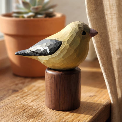 A hand-carved wooden Female American Goldfinch figurine with a pale yellow body, black and white wings, and a pink beak, mounted on a dark brown wooden cylinder stand. The figurine is placed on a rustic wooden windowsill next to a beige curtain, with a potted succulent in the background.