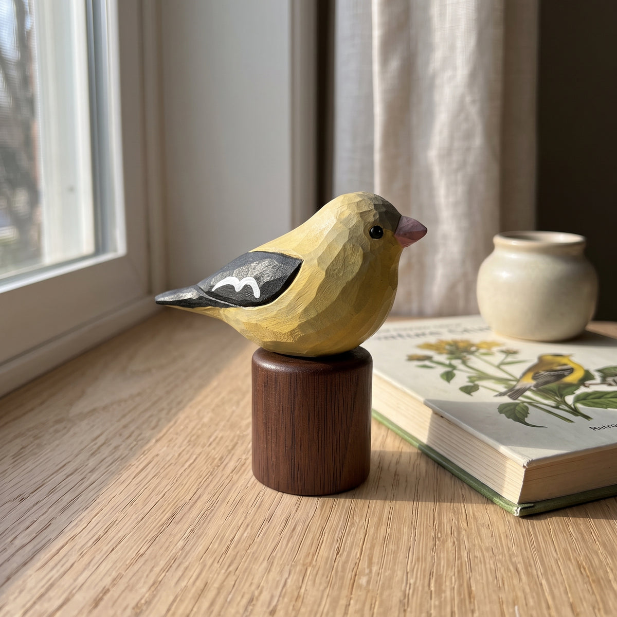 A carved wooden Female American Goldfinch figurine in yellow, black, and gray sits on a dark wood cylindrical base on a sunlit wooden windowsill, with an open book and small vase nearby.