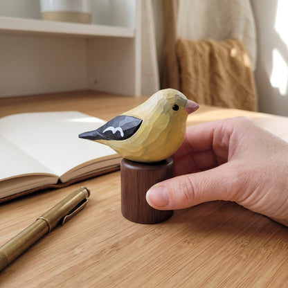 A hand holding a small wooden Female American Goldfinch figurine perched on a cylindrical wooden base, set on a wooden desk with an open book and pen in the background.