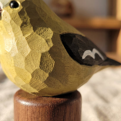 A close-up of a carved wooden Female American Goldfinch figurine with a textured, faceted finish in mustard yellow, black, and white, mounted on a round wooden base.