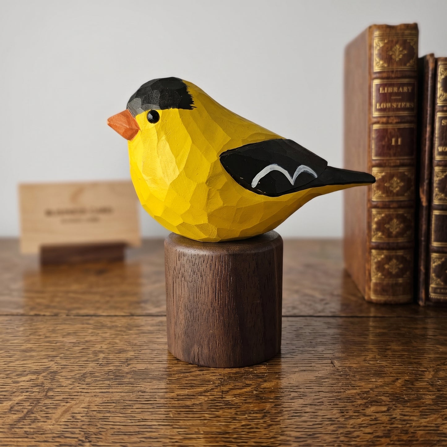 Yellow American Goldfinch figurine on a wooden stand with books in the background
