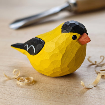 A hand-carved wooden American Goldfinch figurine in bright yellow with black wings and a small orange beak, resting on a light wood surface with carved wood shavings and a carving tool blurred in the background.