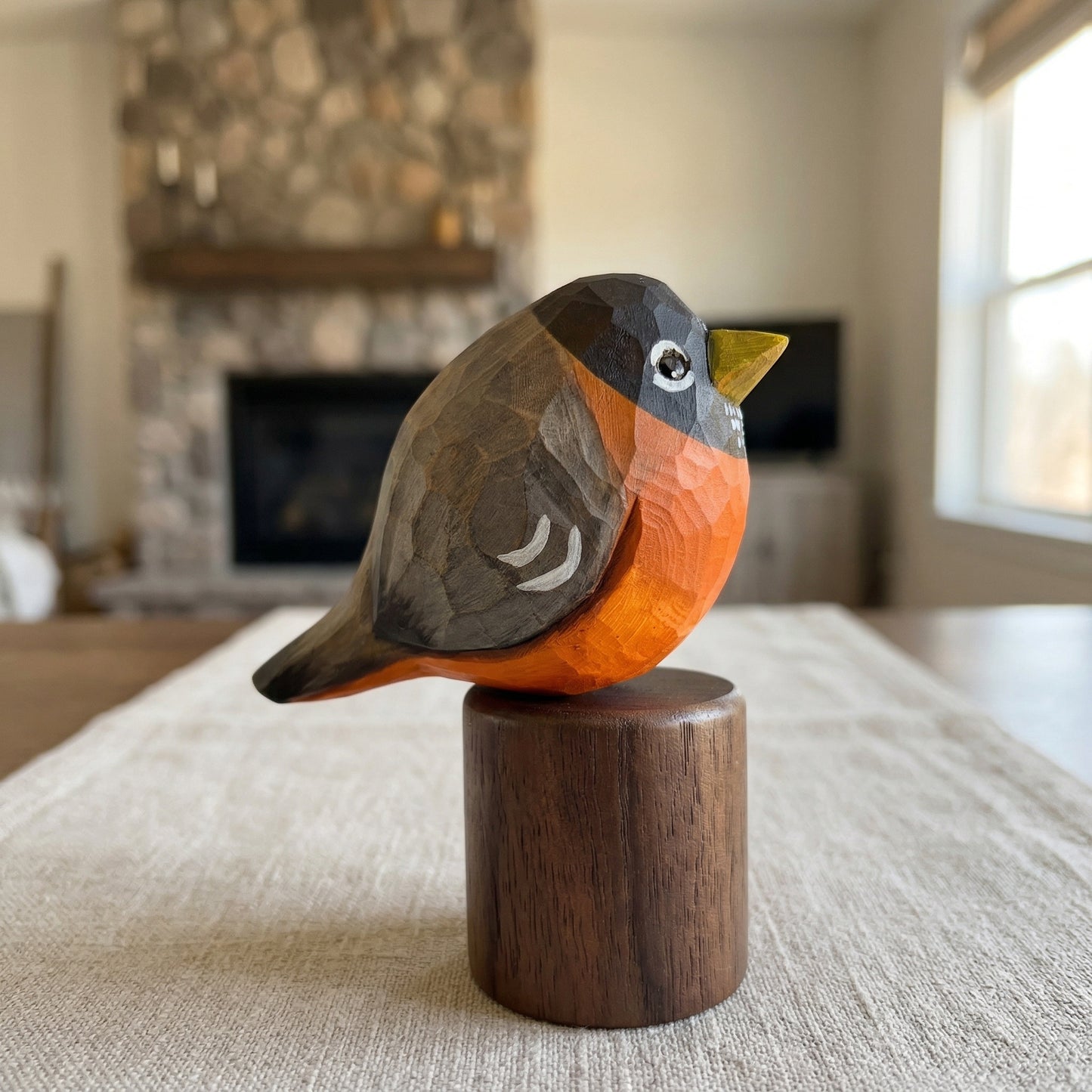 A hand-carved wooden American Robin sculpture with a bright orange chest, dark gray head and back, and a yellow beak, mounted on a round dark wood pedestal. The sculpture is displayed on a beige table runner in a softly blurred living room with a stone fireplace in the background.