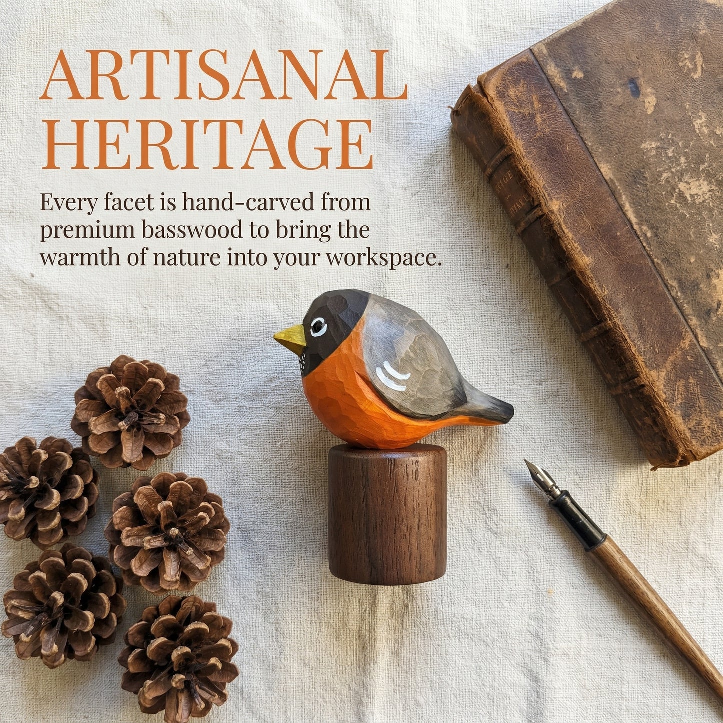 A hand-carved wooden American Robin figurine with an orange breast, dark gray head and wings, and a yellow beak, mounted on a cylindrical wooden base. The bird is styled on a textured off-white fabric surface alongside pinecones, a vintage book, and a fountain pen, with the text 'ARTISANAL HERITAGE' in the upper left.