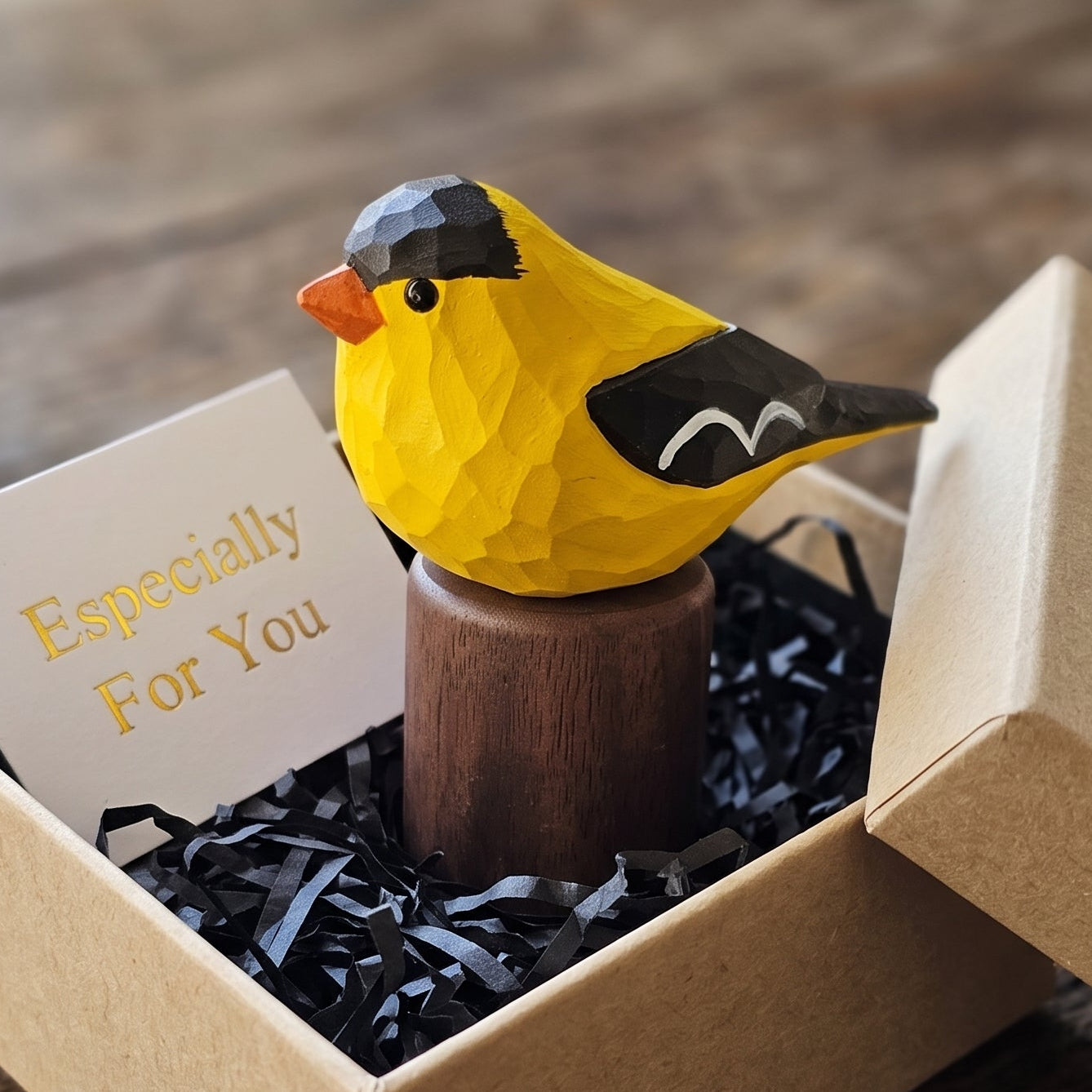 A small yellow American Goldfinch figurine with black and orange details sits on a wooden stand inside an open kraft gift box with shredded black paper filler and a card that reads 'Especially For You'.