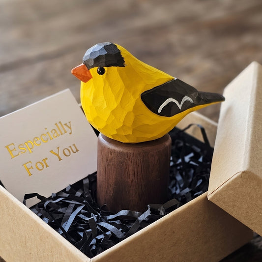 A small yellow American Goldfinch figurine with black and orange details sits on a wooden stand inside an open kraft gift box with shredded black paper filler and a card that reads 'Especially For You'.