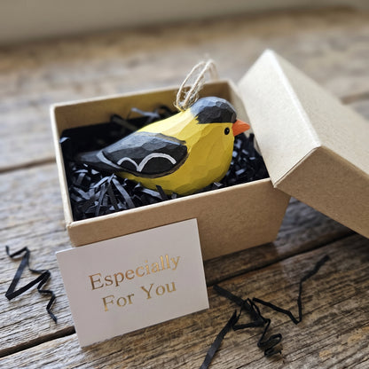 Hand-carved wooden American Goldfinch ornament inside a craft paper gift box with a card