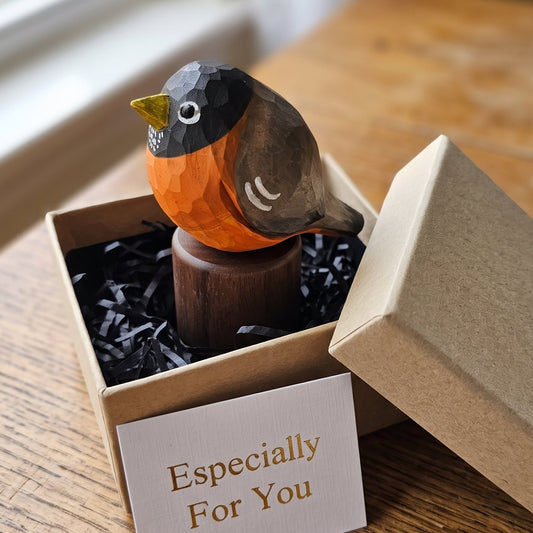 A small carved American Robin figurine with a dark gray head and back, bright orange chest, and yellow beak, displayed in a kraft gift box with black crinkle paper and a card that reads 'Especially For You.' The box sits on a wooden table near a window.