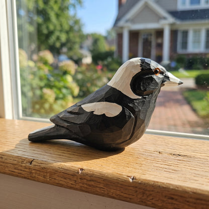 A hand-carved Australian magpie figurine with a black, white, and beige pattern, placed on a wooden windowsill with a blurred outdoor background.