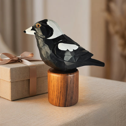 A handcrafted wooden Australian Magpie figurine with a geometric, faceted design, painted in black, white, and gray, perched on a cylindrical wooden base. The figurine is styled on a beige fabric surface next to a small beige gift box with a brown ribbon.