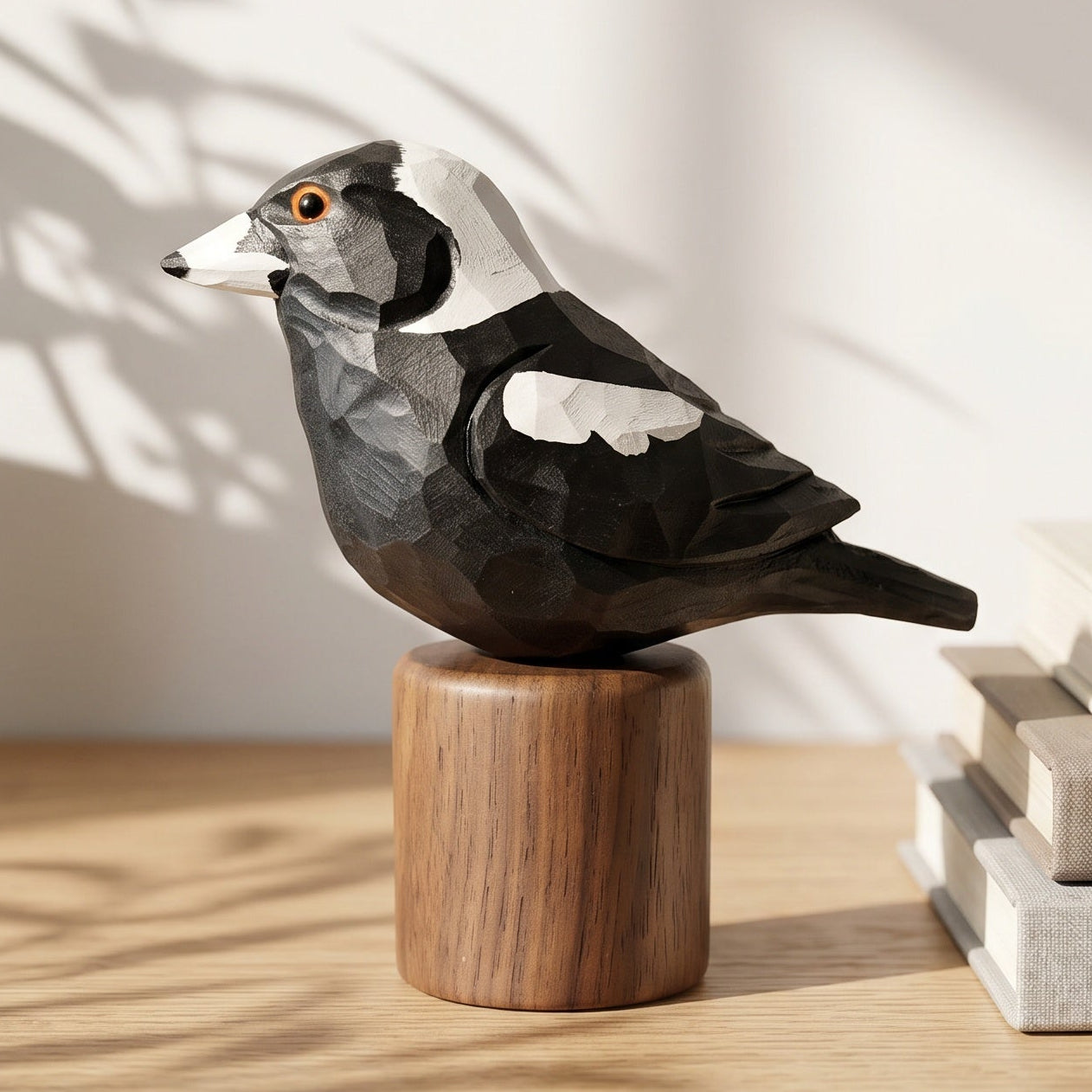 A handcrafted wooden Australian magpie figurine with black, white, and gray geometric facets, perched on a cylindrical wooden base. The figurine is styled on a light wood shelf next to a potted plant and a stack of neutral-colored books, with soft shadows cast across the background.