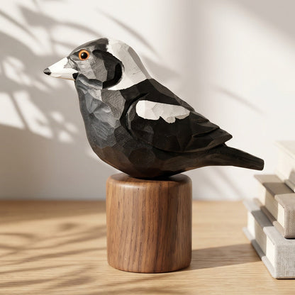 A handcrafted wooden Australian magpie figurine with black, white, and gray geometric facets, perched on a cylindrical wooden base. The figurine is styled on a light wood shelf next to a potted plant and a stack of neutral-colored books, with soft shadows cast across the background.