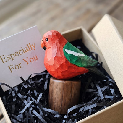 A small carved Australian King Parrot figurine with a bright red-orange body, green wing, and dark eye sits on a wooden stand inside a kraft gift box filled with black shredded paper. A card reading 'Especially For You' is tucked behind it.