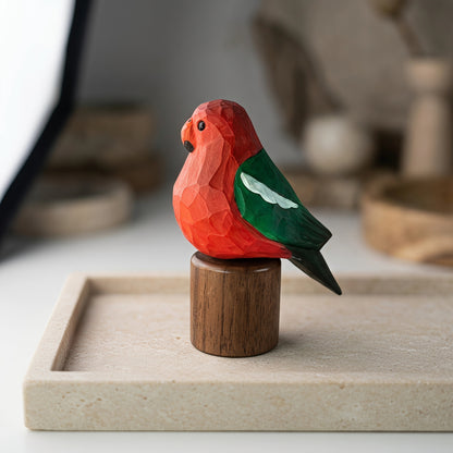 A small carved  Australian King Parrot figurine with a bright red-orange body, green wing, and dark tail, mounted on a cylindrical wooden base and photographed on a light stone tray in a softly blurred indoor setting.