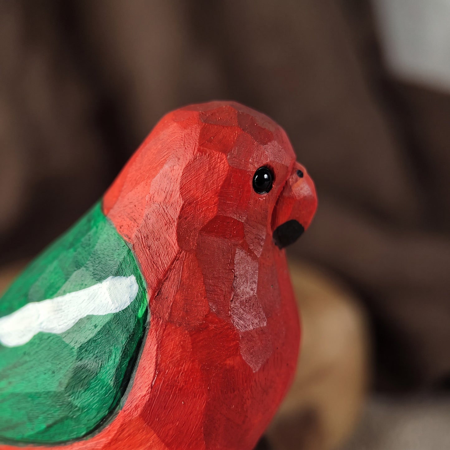A close-up of a hand-carved wooden Australian King Parrot figurine, likely a parrot, featuring a textured red body, a painted green wing, and a small black beak and eye, set against a soft, blurred brown background.