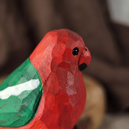 A close-up of a hand-carved wooden Australian King Parrot figurine, likely a parrot, featuring a textured red body, a painted green wing, and a small black beak and eye, set against a soft, blurred brown background.