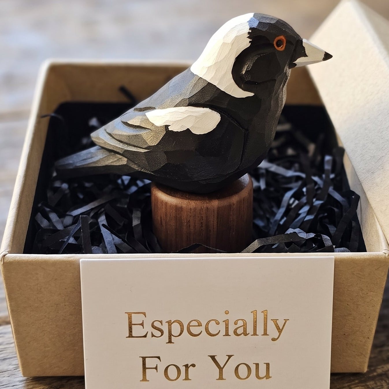 A carved Australian magpie figurine with a black and white body and orange eye, displayed inside a kraft gift box with black shredded paper and a card that reads 'Especially For You'.