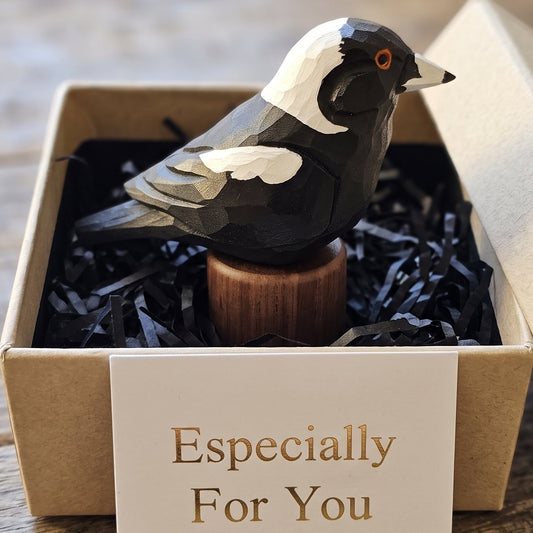 A carved Australian magpie figurine with a black and white body and orange eye, displayed inside a kraft gift box with black shredded paper and a card that reads 'Especially For You'.