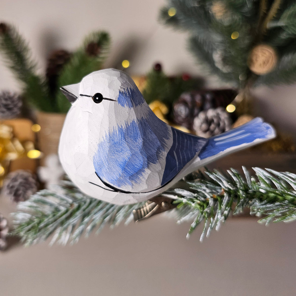 A close-up shot of a hand-carved wooden Azure Tit clip-on ornament perched on a frosted evergreen pine branch, featuring a bright azure-blue back and wings, a pure white head and belly, a black eye-stripe, and warm holiday lights in the background.
