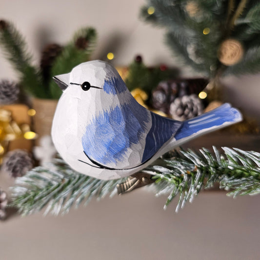 A close-up shot of a hand-carved wooden Azure Tit clip-on ornament perched on a frosted evergreen pine branch, featuring a bright azure-blue back and wings, a pure white head and belly, a black eye-stripe, and warm holiday lights in the background.