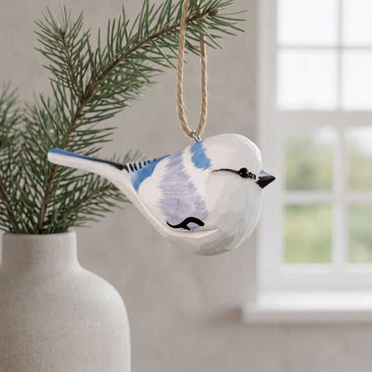 A hand-painted Azure tit Christmas ornament hanging from a pine branch with a rope loop, set against a softly blurred indoor background with a window and a vase.