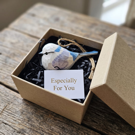 A small decorative Azure tit ornament with a geometric, low-poly style in blue, white, and gray, nestled in black shredded paper inside a brown kraft gift box. A white card reading 'Especially For You' is placed in front.