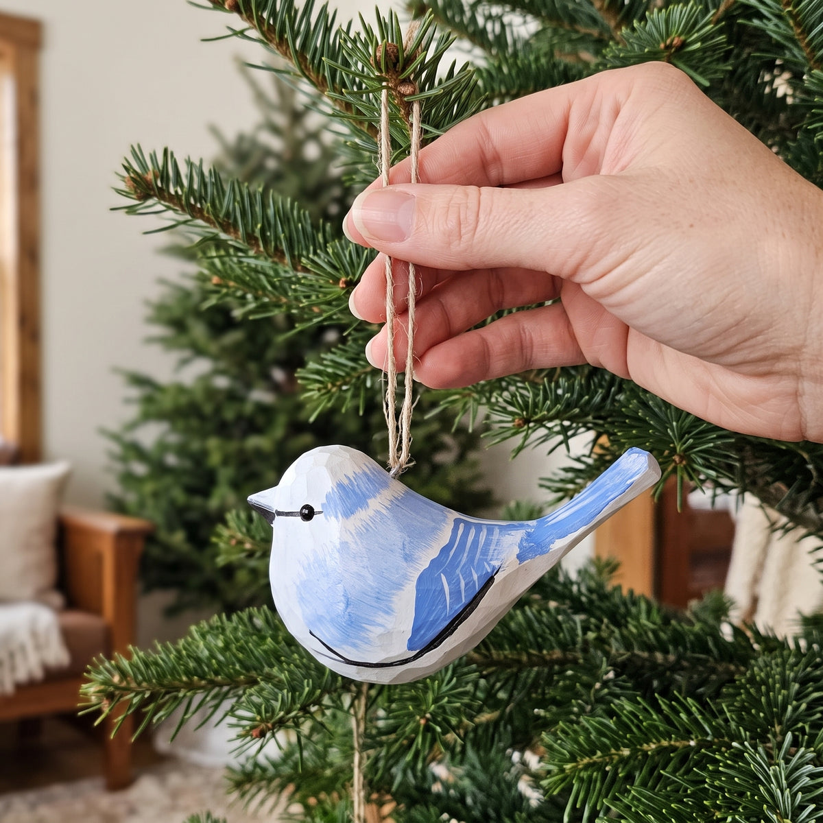 A hand hanging a small painted Azure tit ornament from a pine tree branch in a cozy indoor setting.