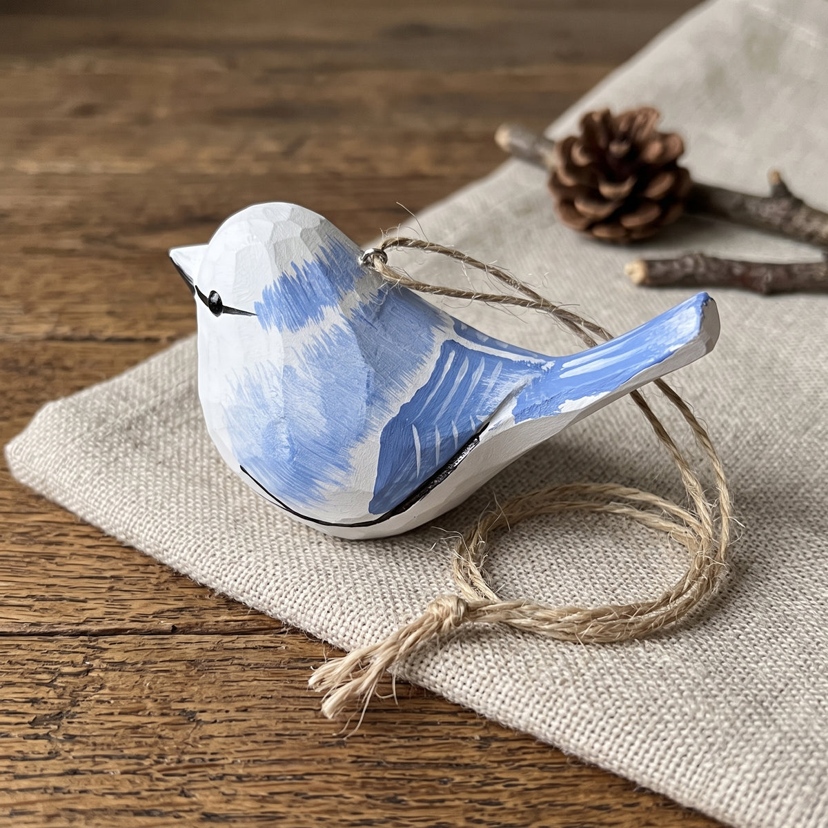 Hand-painted wooden Azure tit ornament in blue and white with a twine loop, displayed on a beige linen cloth over a rustic wooden surface.