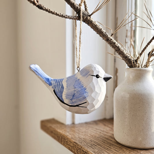 A faceted wooden Azure tit ornament painted in white and light blue, hanging from a branch next to a ceramic vase on a wooden shelf.