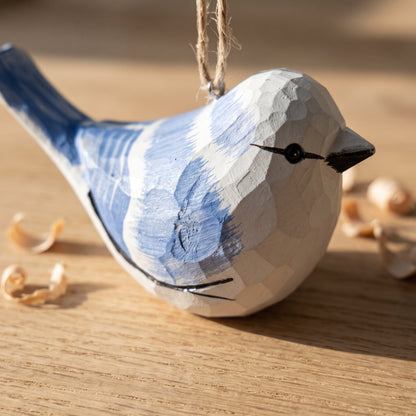 A hand-carved wooden Azure tit ornament painted in blue and white, hanging by a twine loop on a wooden surface with small wood shavings nearby.