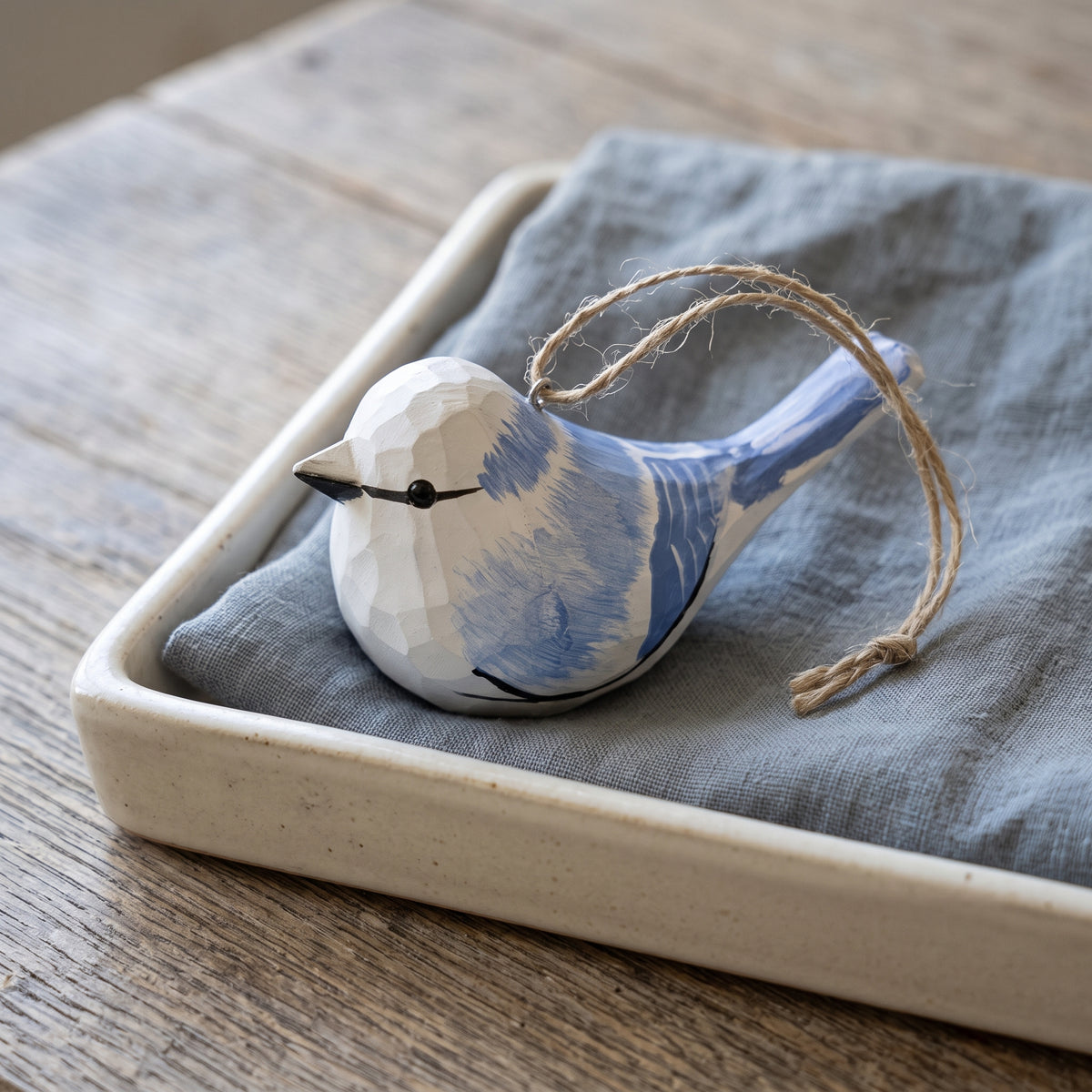 A faceted ceramic Azure tit ornament painted blue and white, with a small black beak and eye, hanging from a natural jute string. The ornament rests on a gray linen cloth inside a square beige ceramic tray, set on a rustic wooden table.