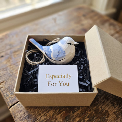 A small wooden Azure tit  ornament with blue and white painted details sits in a brown gift box filled with black shredded paper and a card that reads 'Especially For You'.