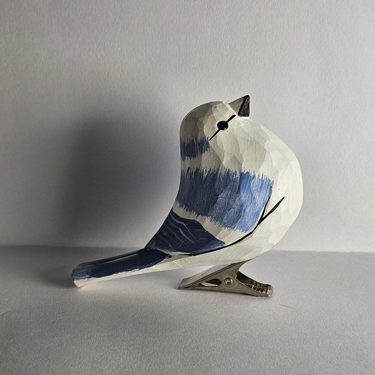 A side-profile shot of a hand-carved wooden Azure Tit clip-on ornament perched upright on a flat surface against a plain light grey background, featuring a white body, bright blue wing and tail accents with a white wing bar, a black eye-stripe, and a silver metal clip attached underneath.