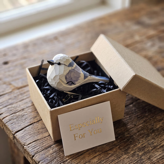 A hand-carved wooden Azure Tit ornament resting in an open kraft gift box filled with black shredded paper, featuring a white head, light gray wing accents, and a small card reading "Especially For You" placed in the foreground.