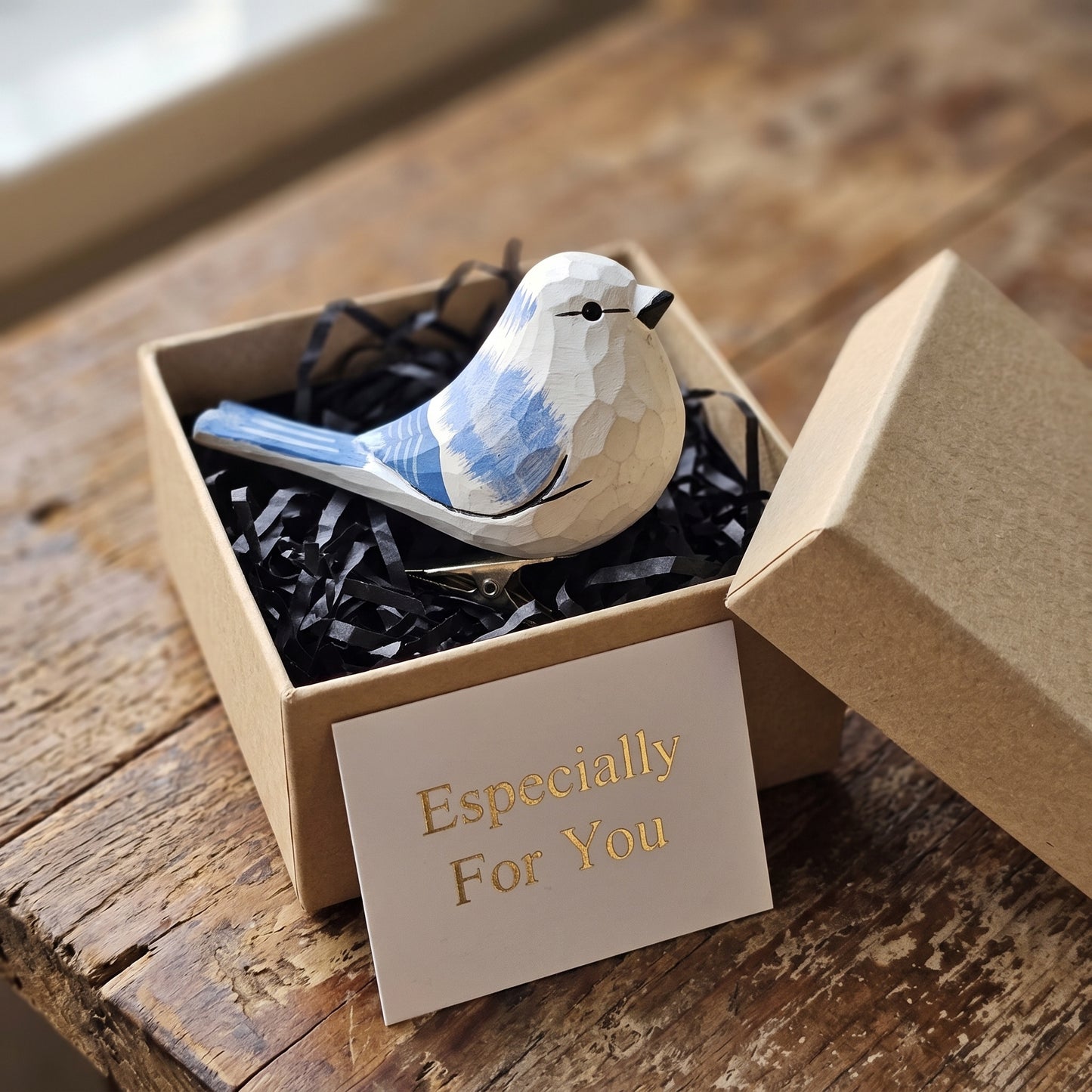 A close-up shot of a hand-carved wooden Azure Tit clip-on ornament resting inside an open kraft gift box filled with black shredded paper, featuring a white head, a blue wing with light-coloured markings, and an gold-foil stamped card reading "Especially For You" placed in the foreground.