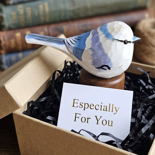 A small painted Azure tit figurine perched on a wooden stand, presented in an open kraft gift box with black shredded paper and a card reading 'Especially For You.' Stacked books and a spool of twine sit blurred in the background on a wooden surface.