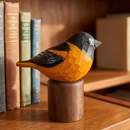 A carved wooden Baltimore Oriole figurine with a bright orange body, black wings, and a gray head, perched on a cylindrical wooden base on a wooden bookshelf beside vintage books.