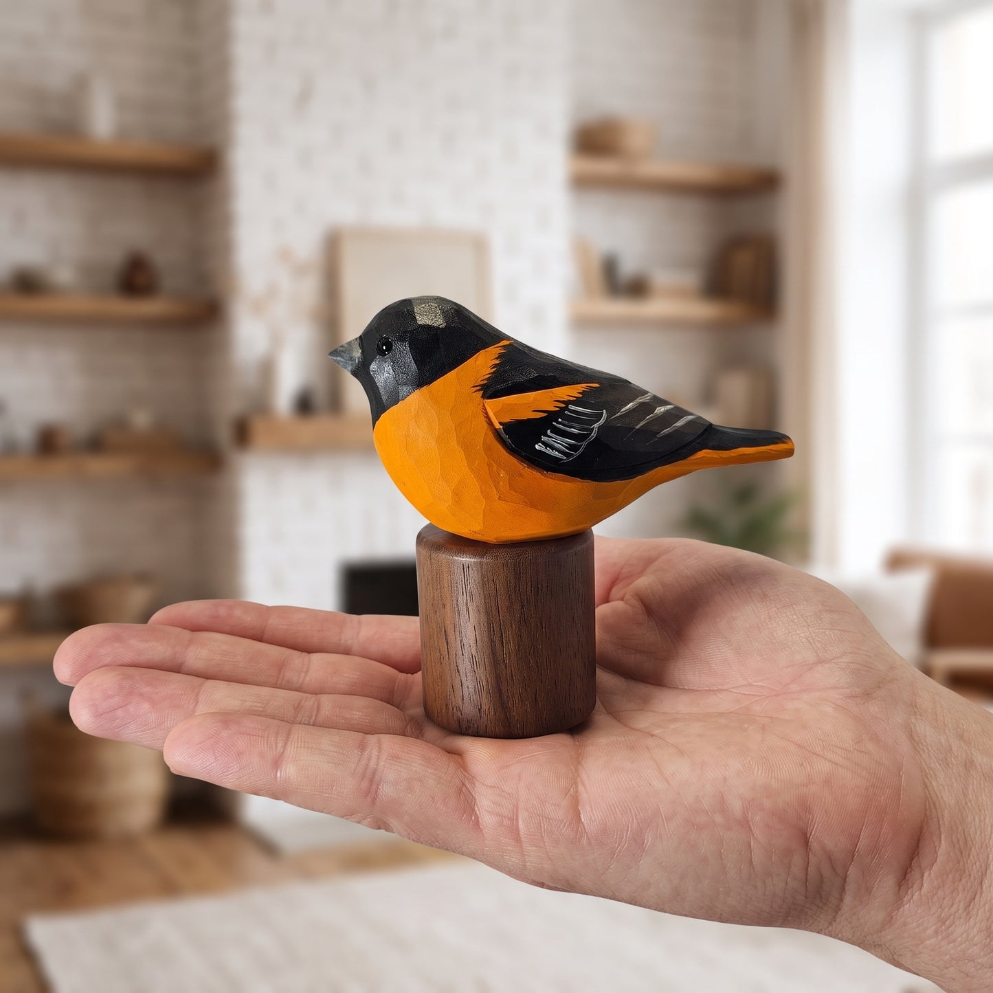 A hand holds a small carved wooden Baltimore Oriole figurine with a bright orange body, black head and wings, and a dark brown wooden base, set against a softly blurred living room background.