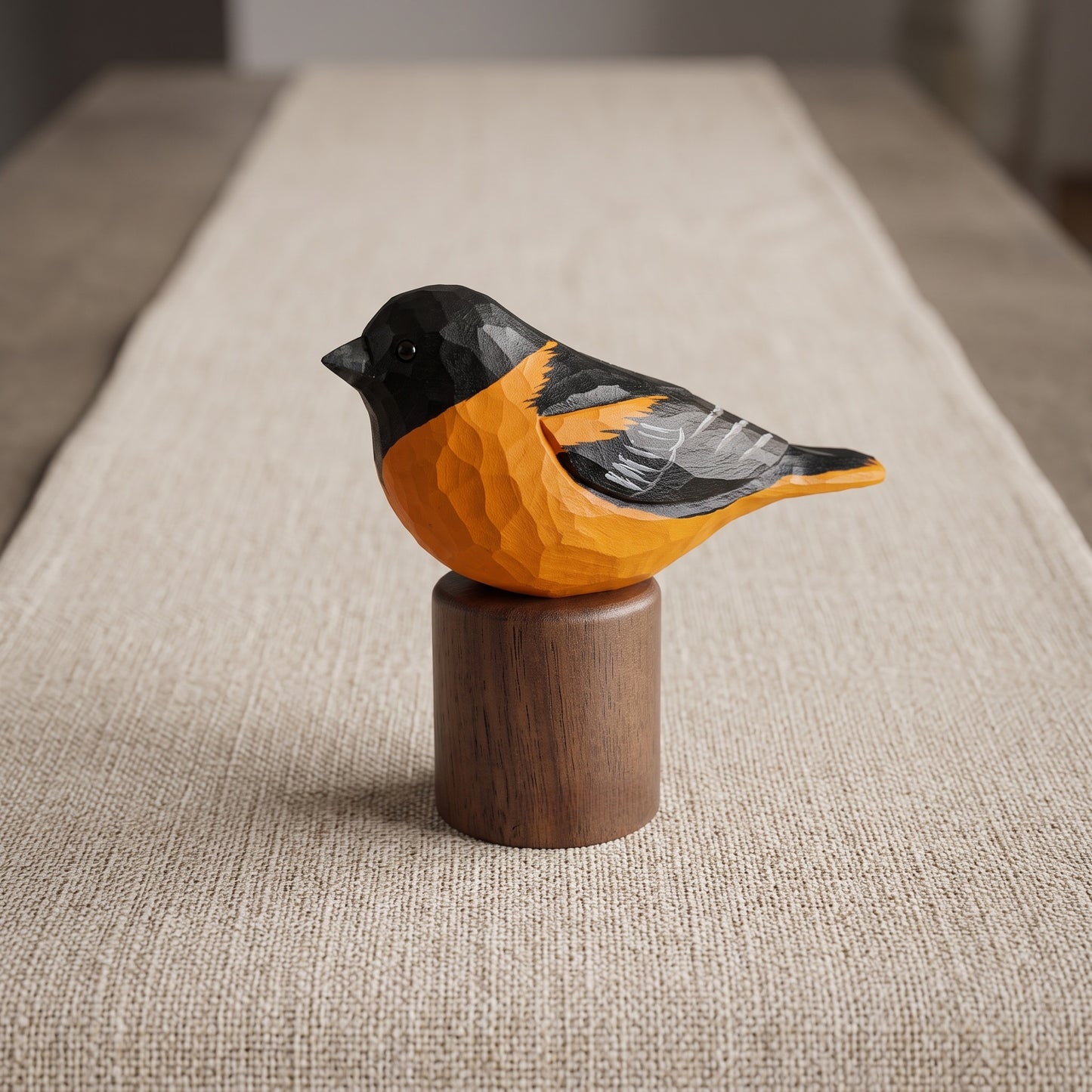 A carved wooden Baltimore Oriole sculpture with a bright orange body, black head and back, and gray wing details, perched on a cylindrical dark wood base over a beige woven runner.