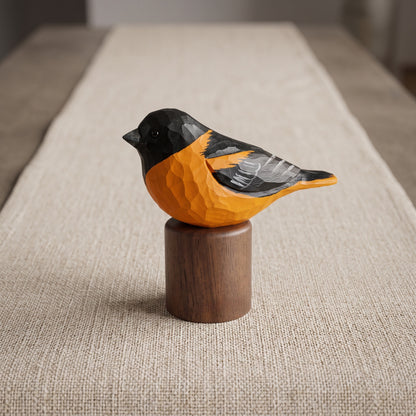 A carved wooden Baltimore Oriole sculpture with a bright orange body, black head and back, and gray wing details, perched on a cylindrical dark wood base over a beige woven runner.