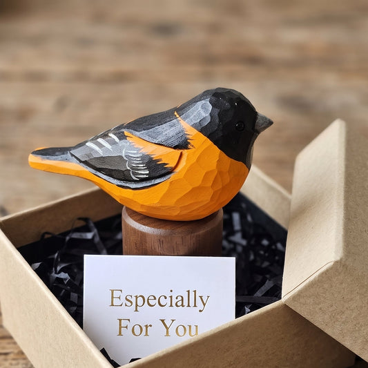 A hand-carved wooden Baltimore Oriole figurine with a bright orange body, black head, and gray wings, displayed in an open kraft gift box with black paper filler and a card that reads 'Especially For You' on a rustic wooden table.