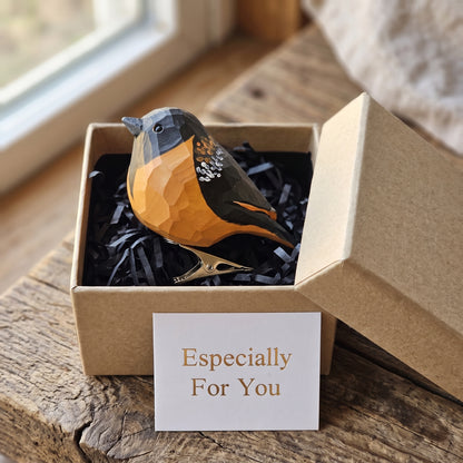 A small Baltimore Oriole clip-on-shaped decorative item in a brown kraft gift box with black shredded paper, placed on a rustic wooden surface near a window. A white card in front reads 'Especially For You'.
